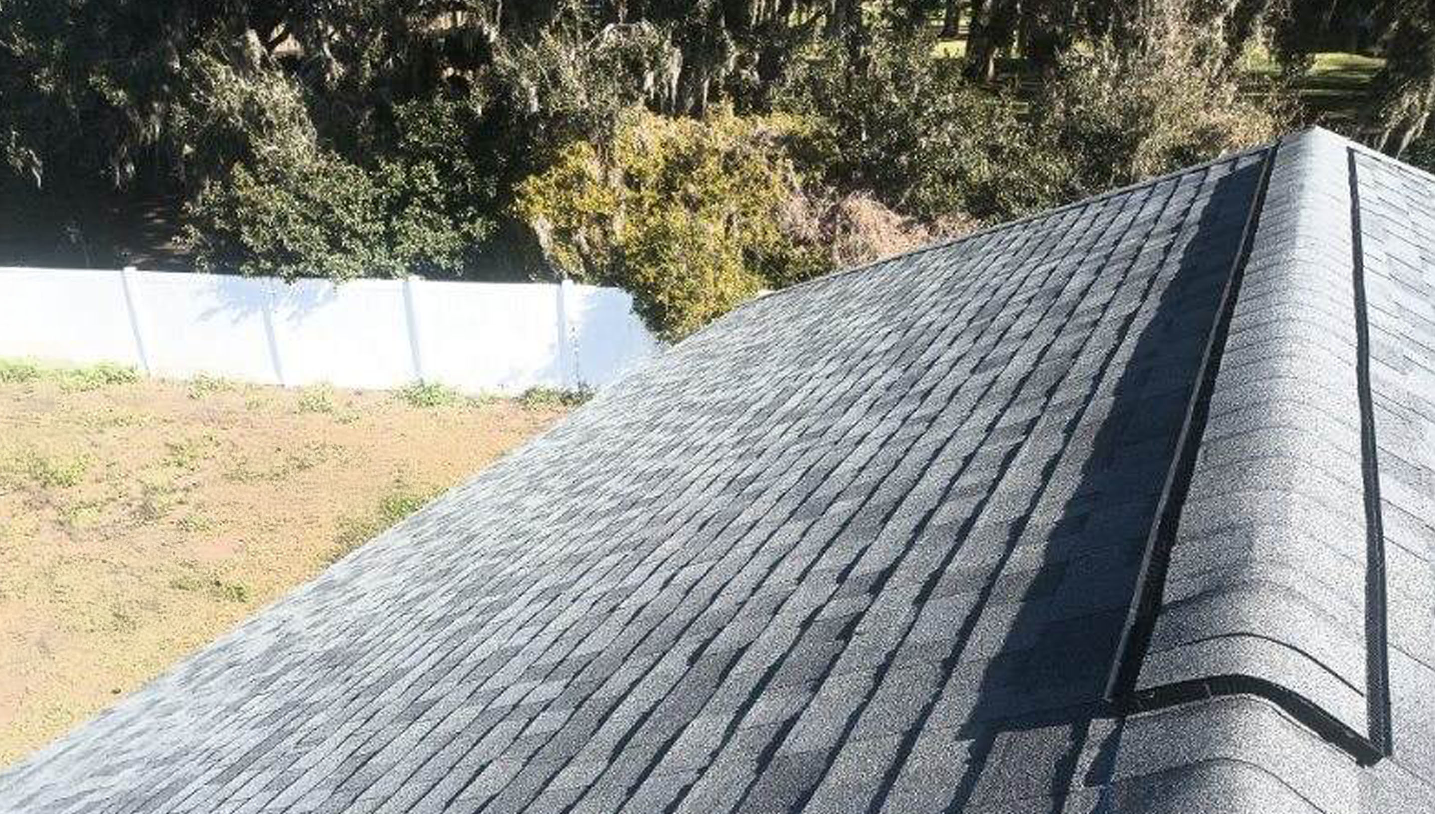 A rooftop with grey asphalt shingles, a white fence, and trees in the background.